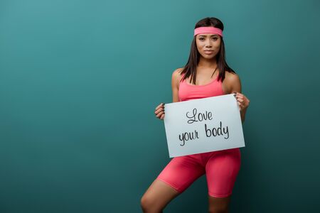 African american sportswoman smiling and looking at camera with placard with love your body lettering isolated on greenの写真素材