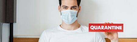 Man in medical mask holding card with quarantine lettering at home, panoramic crop