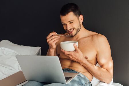 happy and shirtless man holding bowl with spoon and watching movie on laptop
