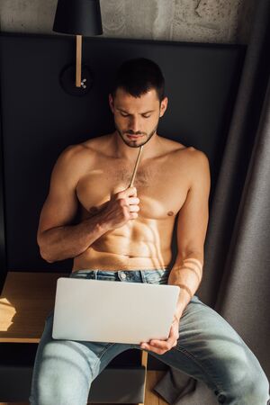 bearded and shirtless freelancer holding pencil and looking at laptopの写真素材