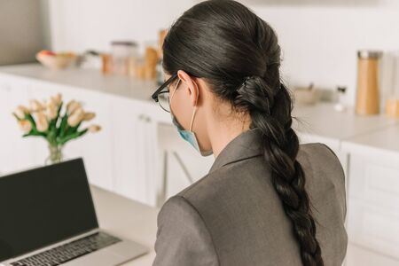 brunette, long haired businesswoman in medical mask working on laptop in kitchen