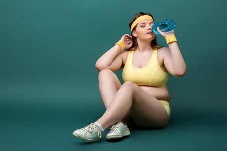 Plus size sportswoman sitting with crossed legs and drinking water on green backgroundの写真素材