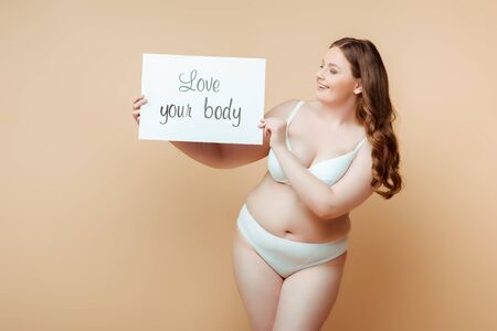 Plus size woman smiling and showing placard with love your body lettering on beigeの写真素材