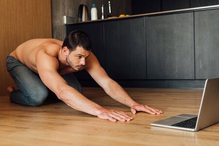 muscular man doing balasana exercise while watching online yoga training on laptopの写真素材