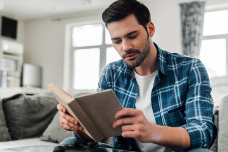 Selective focus of handsome man in plaited shirt reading book at homeの写真素材
