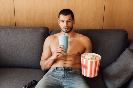 shirtless man holding popcorn bucket and drinking soda near remote controller on sofa