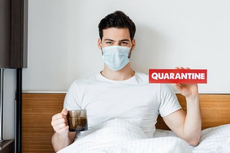 Man in medical mask holding card with quarantine lettering and cup of coffee on bedの写真素材