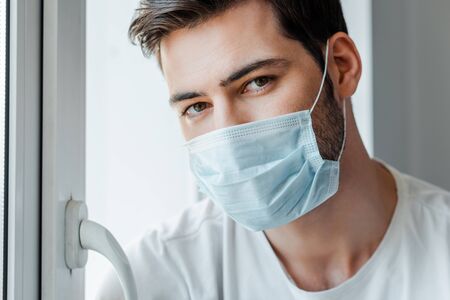 Man in medical mask looking at camera near window at homeの写真素材