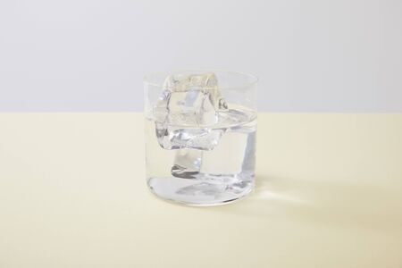 Glass with water and ice cubes on yellow surface isolated on grey