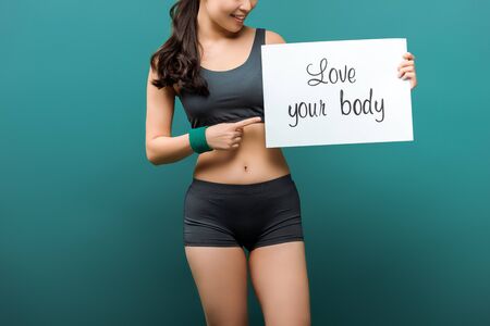 Cropped view of sportswoman smiling and pointing at placard with love your body lettering on green backgroundの写真素材