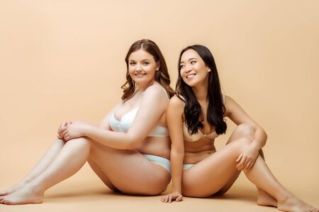 cheerful overweight woman and happy asian girl in underwear sitting on beige, body positive conceptの写真素材