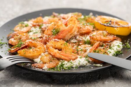 selective focus of fried shrimps in sauce with dill and lemon in black plate on grey concrete background with cutleryの写真素材
