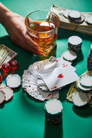 cropped view of woman holding glass of cognac near money, playing cards, dice and casino tokens on...