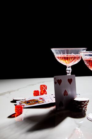 playing cards, dice and casino chips near glasses of cocktail on white surface isolated on black