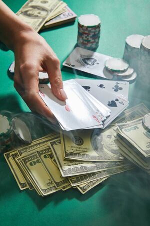 cropped view of woman holding playing cards near dollar banknotes and casino tokens with smoke...