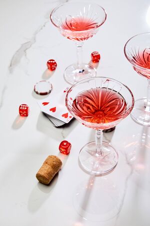high angle view of deck of cards, glasses of cocktail, dice, casino token and cork on white...