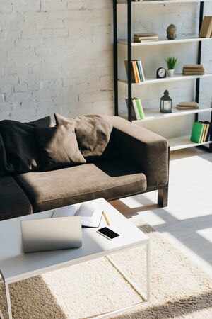 living room with grey sofa, shelf and table with laptop, smartphone and notepad in sunlightの写真素材