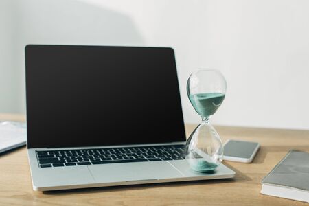 Hourglass on laptop with blank screen near smartphone and notebook on table in officeの写真素材