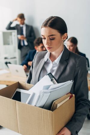 Selective focus of fired woman holding box with documents and digital tablet while colleagues working in officeの写真素材