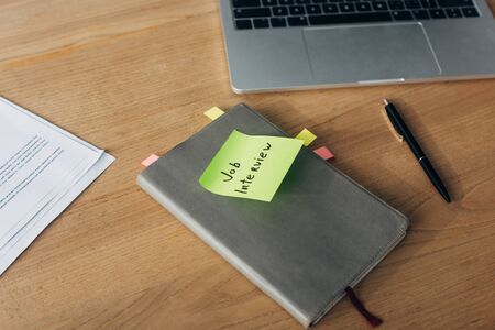 Notebook with job interview lettering on sticky note near laptop and documents on tableの写真素材