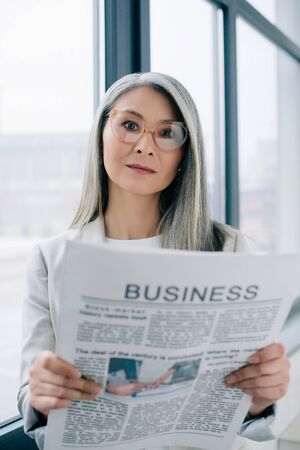 confident asian businesswoman in eyeglasses holding business newspaper in office