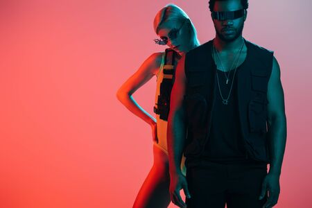 stylish interracial couple posing in futuristic look and sunglasses on pinkの写真素材
