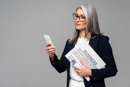 confident asian businesswoman in eyeglasses using smartphone and holding business newspaper isolated on greyの写真素材