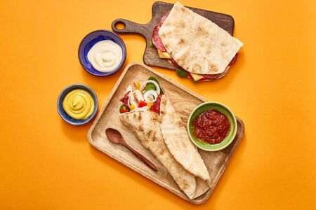 top view of fresh sandwich with salami in pita and burrito with chicken on boards near sauces on orange backgroundの写真素材