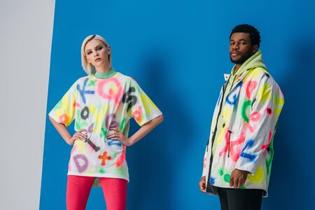 fashionable multiethnic couple posing in colorful futuristic look on grey and blue