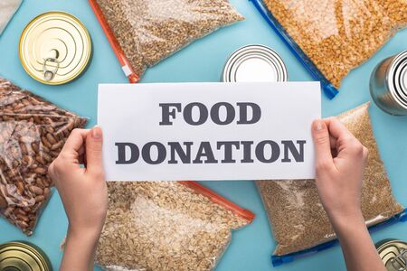 cropped view of woman holding card with food donation lettering near cans and groats in zipper bags on blue backgroundの写真素材