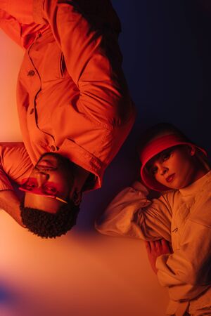 top view of fashionable interracial couple in futuristic clothes lying in orange light
