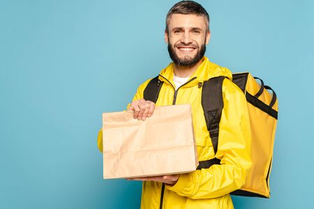 smiling deliveryman in yellow uniform with backpack holding paper package on blue backgroundの写真素材