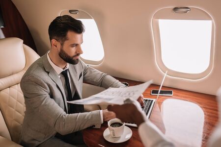 cropped view of african american stewardess giving newspaper to businessman working on laptop in...