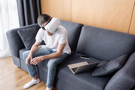 Man in vr headset and medical mask sitting near smartphone and laptop on couch