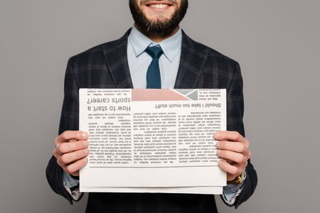 cropped view of smiling bearded businessman in suit with newspaper on grey backgroundの写真素材