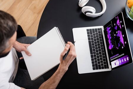 Overhead view of freelancer holding notebook near laptop with charts on screen