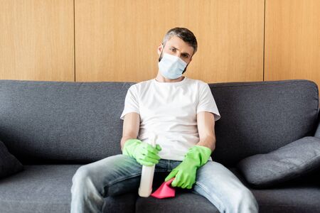 Man in medical mask holding rag and detergent while sitting on couchの写真素材