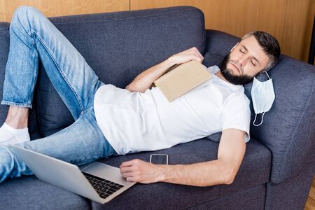 Freelancer in medical mask sleeping near book and digital devices on couch