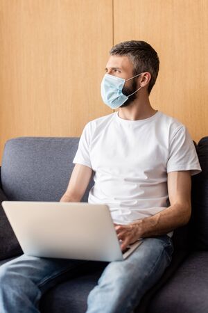 Selective focus of teleworker in medical mask looking away while using laptop at homeの写真素材