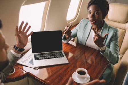 angry african american businesswoman showing middle fingers while sitting opposite businessman in private jetの写真素材
