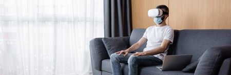 Panoramic shot of man in medical mask and vr headset sitting near laptop on couchの写真素材