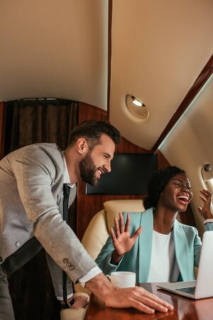 smiling businessman standing near excited african american businesswoman waving hand while having...