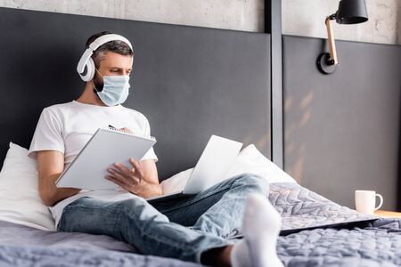 Selective focus of teleworker in headphones and medical mask whiting on notebook and using laptop in bedroomの写真素材