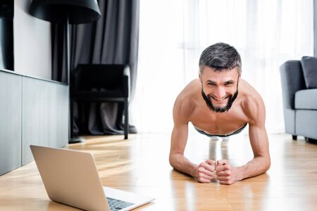 Shirtless man smiling at camera while standing in plank near laptop on floor at home