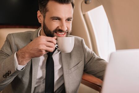smiling businessman drinking coffee and looking at laptop in private plane