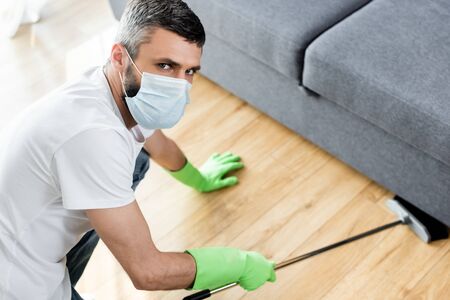 Man in medical mask looking at camera while cleaning floor in living room