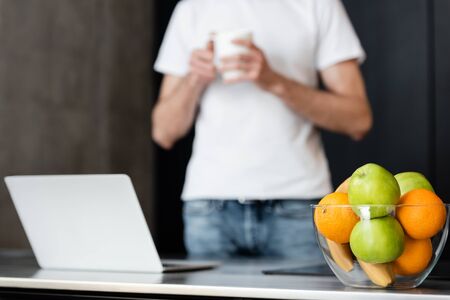 Selective focus of fresh fruits in bowl and man holding cup near laptop on kitchen worktop