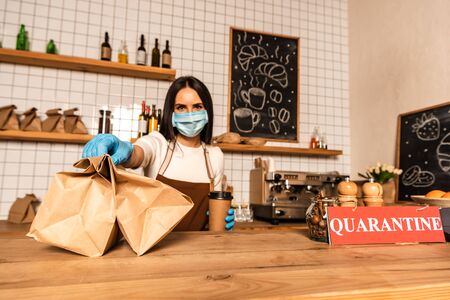 Cafe owner in medical mask with disposable cup of coffee showing paper bags near table with card with closed inscriptionの写真素材