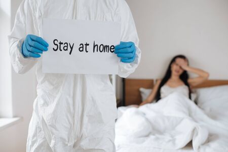 cropped view of doctor in protective suit holding card with Stay at home lettering in bedroom with ill woman, selective focusの写真素材