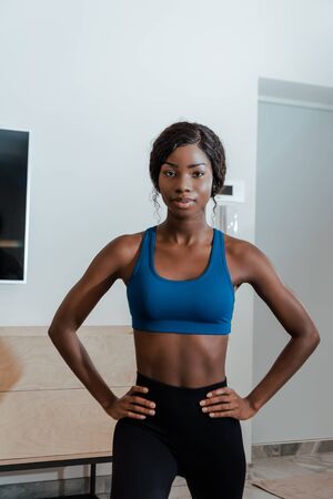 Front view of african american sportswoman with hands on hips doing lunges and looking at camera in living roomの写真素材